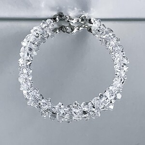 May include: A clear, circular bracelet composed of numerous small, irregularly shaped, clear crystals. The bracelet is connected by a silver chain. The crystals reflect light, creating a sparkling effect. The jewelry is set against a light gray background.
