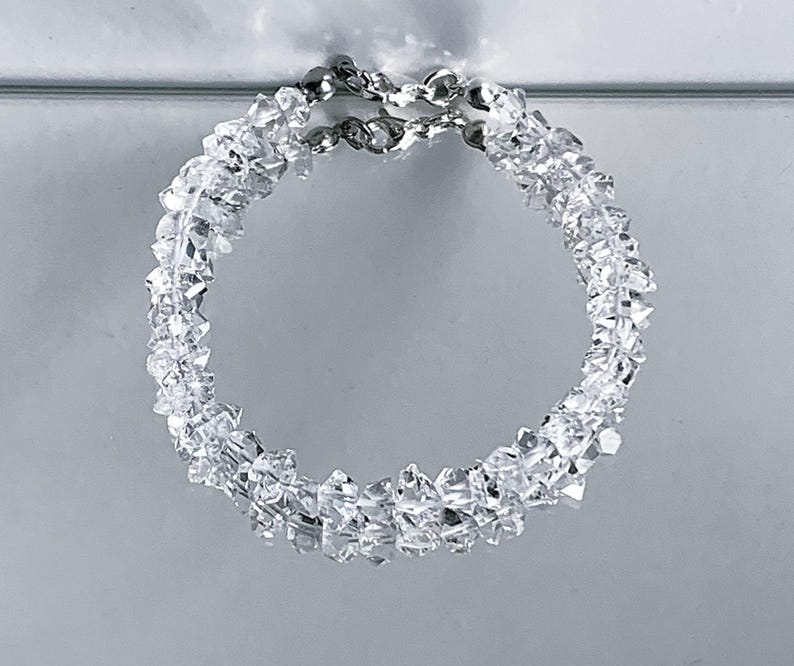 May include: A clear, circular bracelet composed of numerous small, irregularly shaped, clear crystals. The bracelet is connected by a silver chain. The crystals reflect light, creating a sparkling effect. The jewelry is set against a light gray background.