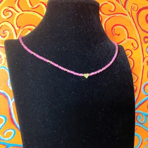 May include: A pink beaded necklace with a gold heart charm. The necklace is displayed on a black velvet mannequin bust.