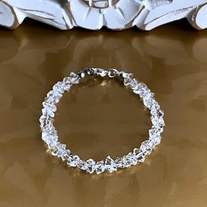 May include: A clear quartz crystal bracelet. The bracelet is made of small, irregularly shaped quartz crystals strung together with a silver-colored clasp. The crystals are translucent and reflect light, creating a sparkling effect. The bracelet is a delicate piece of jewelry.