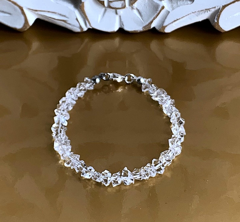 May include: A clear quartz crystal bracelet. The bracelet is made of small, irregularly shaped quartz crystals strung together with a silver-colored clasp. The crystals are translucent and reflect light, creating a sparkling effect. The bracelet is a delicate piece of jewelry.