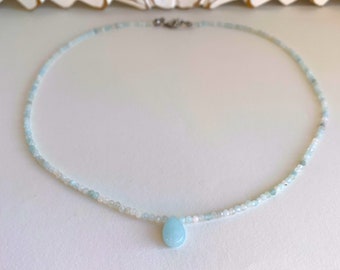 May include: A delicate necklace made of light blue and white beads with a single teardrop-shaped light blue gemstone pendant.