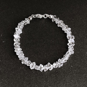 May include: A clear crystal bracelet with a silver clasp. The bracelet is composed of irregularly shaped, faceted crystals strung together. The crystals are transparent and reflect light, creating a sparkling effect. The bracelet is shown against a dark background.