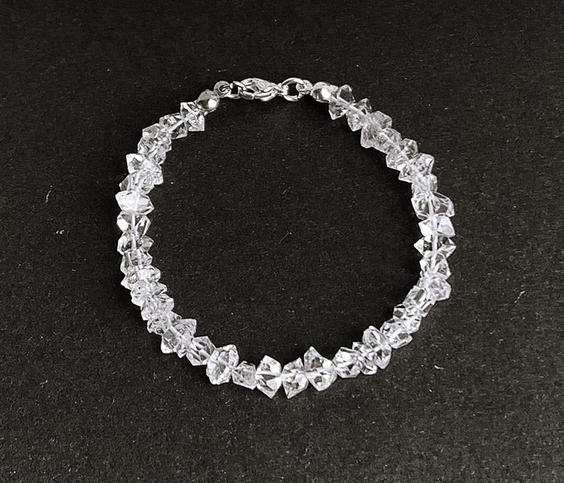May include: A clear crystal bracelet with a silver clasp. The bracelet is composed of irregularly shaped, faceted crystals strung together. The crystals are transparent and reflect light, creating a sparkling effect. The bracelet is shown against a dark background.