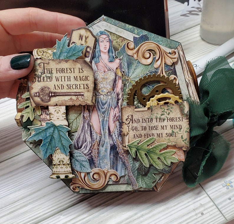 Puede incluir: Una p&aacute;gina de &aacute;lbum de recortes hexagonal con un esquema de color verde, marr&oacute;n y dorado. La p&aacute;gina presenta a una mujer con cabello largo y suelto y un vestido azul, rodeada de hojas, flores y una cita que dice "El bosque est&aacute; lleno de magia y secretos". La cita "Y al bosque voy, a perder mi mente y encontrar mi alma" tambi&eacute;n aparece en la p&aacute;gina.