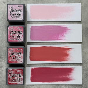 May include: Four Distress Oxide ink pads with color swatches. The ink pads are labeled "spun sugar", "picked raspberry", "festive berries", and "lumberjack plaid". Each swatch shows a brush stroke of the corresponding color on a white card.