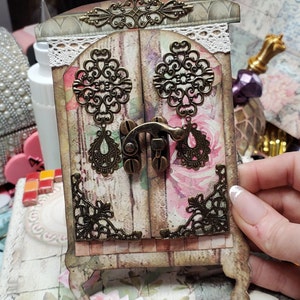 Puede incluir: Un pequeño gabinete de madera decorativo con una puerta con bisagras y un pestillo de metal. El gabinete está pintado en blanco desgastado y presenta diseños florales y detalles metálicos ornamentados. El gabinete está sentado en una mesa con un patrón floral.
