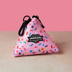 Op de afbeelding: Roze driehoekige tas met een ritssluiting en een zwart hartvormig logo met de tekst "NY Ruffhaus C". De tas heeft een kleurrijk spikkels patroon.