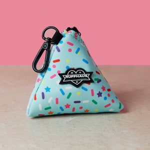 Op de afbeelding: Een lichtblauwe driehoekige tas met een zwarte rits en een zwart hartvormig patch met de tekst "NY Ruffhaus C" erop. De tas heeft een zwarte clip en is bedekt met een kleurrijk sprenkelpatroon.