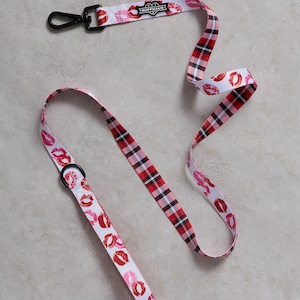 May include: A red and white plaid dog leash with a white background and red lips printed on it. The leash has a black clip and a black tag that says "Ruffhaus" with a heart.