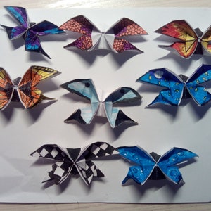Könnte beinhalten: Acht Origami-Schmetterlinge mit verschiedenen farbigen Mustern und Designs. Die Schmetterlinge sind aus Papier und in eine traditionelle Schmetterlingsform gefaltet. Die Schmetterlinge sind in einer Reihe auf einer weißen Oberfläche angeordnet.