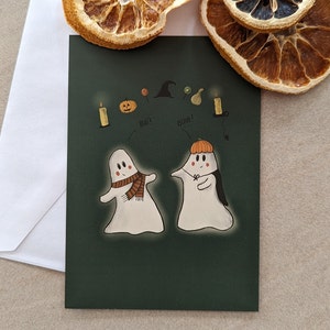 Puede incluir: Una tarjeta verde con dos fantasmas blancos, uno con una bufanda y el otro con un sombrero de calabaza. La tarjeta tiene las palabras "Boo!" y "Happy" escritas en ella. También hay ilustraciones de una vela, una calabaza, una piruleta, un sombrero de bruja, una cebolla verde y un diente de ajo.