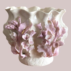Op de afbeelding: Een crèmekleurige keramische vaas met een golvende bovenrand en een getextureerde voet. De buitenkant is versierd met driedimensionale, lichtroze bloem- en bladmotieven met delicate goudkleurige accenten op de bloemblaadjes en bladeren.