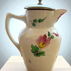 Peut inclure: Une cruche en céramique blanche avec un couvercle et une anse, ornée de motifs floraux roses et jaunes et de feuilles vertes. La cruche a un bouton de couleur laiton sur le couvercle et un bec verseur avec des accents rouges. Un objet décoratif pour servir des boissons.