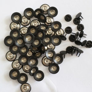 May include: A collection of black metal snap fasteners with a concave center and a silver-toned metallic core. The fasteners feature a circular rim and are arranged in a cluster, with some showing the pronged back side facing upward.
