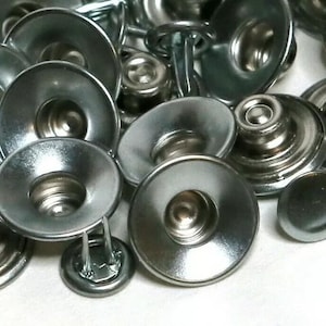 Op de afbeelding: Een close-up van een stapel zilveren metalen drukknopen. De drukknopen hebben een ronde, gewelfde bovenkant en een puntige basis.