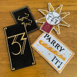 May include: Four embroidered patches on a wooden surface. Two black rectangular patches with gold numbers, one with "33" and the other with a stylized "33". A white patch with a baguette and the text "PARRY IT!". A sun-shaped patch with a face.