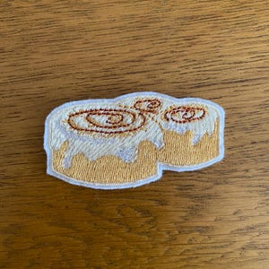 May include: Embroidered patch depicting a cinnamon roll. The patch features white icing with brown swirls and a tan-coloured roll base. The patch is outlined in white, a fun accessory for clothing or bags.
