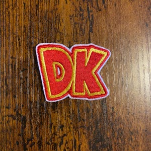 May include: An embroidered patch featuring the letters "DK" in red with a yellow outline and a white border. The patch has a bold, stylized design. It is likely intended for application on clothing or accessories.