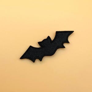 May include: A black embroidered patch in the shape of a bat with outstretched wings. The design features detailed stitching throughout the body and wings, creating a textured surface. The bat is depicted in a flying position with pointed ears.