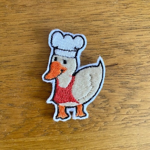 May include: Embroidered patch of a cartoon duck in a chef's hat and apron. The duck has an orange beak and feet, a red apron, and a white chef's hat. The patch is outlined in white and is on a wooden surface.