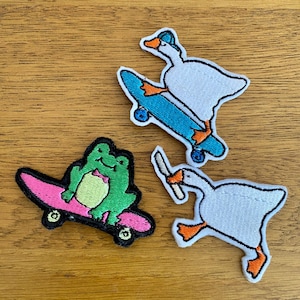 May include: Three embroidered iron-on patches featuring a green frog, a white goose, and a white goose holding a knife, all riding skateboards. The frog is wearing a bow tie and the goose is wearing a blue cap.