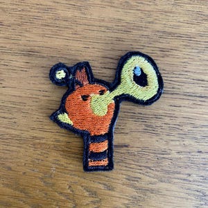 May include: Embroidered patch of a cartoon giraffe playing a yellow trumpet. The giraffe has orange and yellow details with a black outline. Suitable for clothing or accessories.