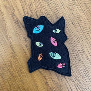 May include: An irregular, black embroidered patch featuring multiple stylised eyes in various colours including light blue, lime green, pink, and orange. The patch has a thick black stitched border and a textured surface created by dense embroidery thread.