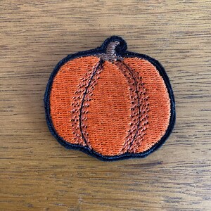 May include: An embroidered orange pumpkin patch with a black border and a brown stem. The pumpkin is stitched with orange thread. This patch is a seasonal item, ideal for autumn and Halloween.