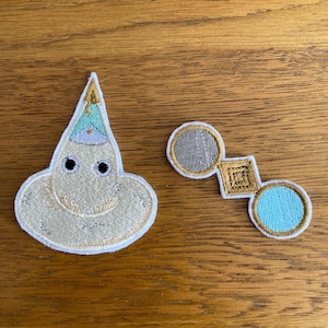May include: Two embroidered patches on a wooden surface. One patch is a cream-coloured, smiling ghost-like figure with a pointed hat in mint green and gold. The other patch is a pair of spectacles with a grey lens and a blue lens.