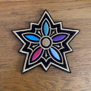 May include: Embroidered patch in a starburst design. The patch has a black background with a gold border. The centre has a gold circle with colourful petals in blue, pink, purple, and teal. Suitable for jackets, bags, or other fabric items.