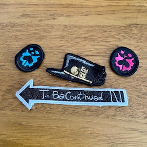 May include: Three black embroidered patches with white and gold stitching. The patches feature a top hat, a gear with a clover, and a gear with a pink clover. A black arrow patch with white stitching reads "To Be Continued".