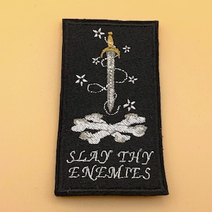 May include: A rectangular black embroidered patch featuring a silver sword with a gold hilt, surrounded by small white stars and a dotted line. Below the sword are two crossed white bones, and the text SLAY THY ENEMIES in white lettering.