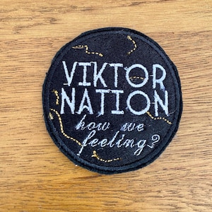 May include: Black round embroidered patch with white text that reads "Viktor Nation how we feeling?" The text is outlined in gold thread.