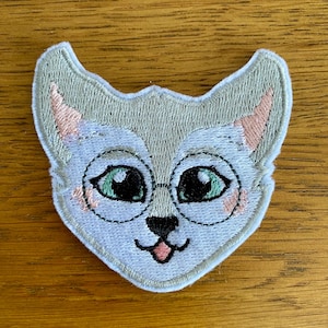 May include: Embroidered patch of a cat's face wearing glasses. The cat has light grey fur, pink ear and cheek details, and green eyes. The design uses black outlines and a light blue background.