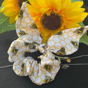 May include: White scrunchie with a bee and honeycomb pattern. The scrunchie is tied in a bow shape. A yellow sunflower and a bee pendant necklace are in the background.