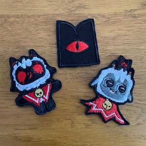 May include: Three embroidered patches on a wooden surface. Two patches depict cartoon lambs in red and black outfits with a skull emblem. The third patch is black with a red eye design. These patches are likely for clothing or accessories.