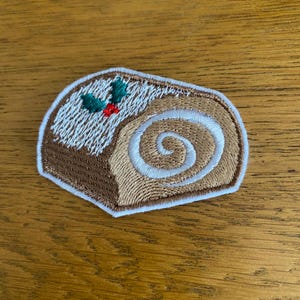 May include: Embroidered patch depicting a slice of a Yule log cake. The cake has a brown exterior, a white swirl interior, and is topped with white frosting, green holly leaves, and red berries. The patch is outlined in a gold colour.