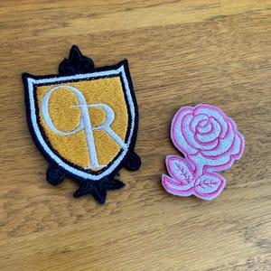 May include: Two embroidered patches on a wooden surface. One is a shield shape with a gold background, white lettering, and a navy blue border. The other is a pink and white rose.