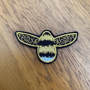 May include: Embroidered bee patch with black and gold detailing. The bee has black stripes on its body and intricate gold patterns on its wings. The patch has a light blue accent at the bottom.