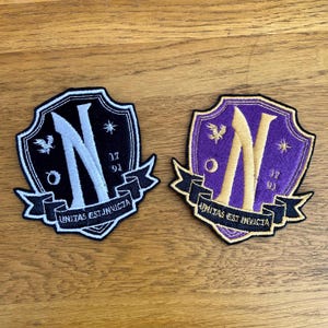 May include: Two embroidered patches with a shield design. One is black with a white "N" and the other is purple with a gold "N". Both have the text "UNITAS EST INVICTA" on a banner.
