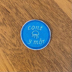 May include: A circular, blue embroidered patch with white edging. The patch displays the words "cont y môr" and a jellyfish design in white thread.