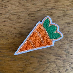 May include: Embroidered carrot patch with orange body and green top. The patch has a white border and is set against a wooden background. Ideal for adding to clothing, bags, or other fabric items.