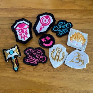 May include: A collection of ten embroidered iron-on patches featuring various designs, including a skull, a smiley face, a shield with an anchor, and a cartoon character. The patches are all different colours and sizes, and they are perfect for adding a unique touch to clothing, bags, or other accessories.