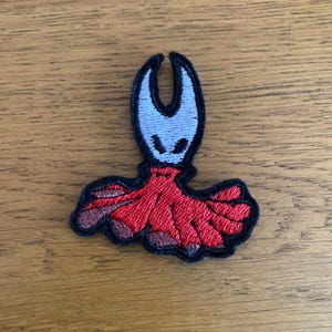 May include: Embroidered patch of a stylized character with a white face, black eyes, and a black outline. The character has a red cape with a black border. The patch is approximately 7.6 cm tall and 5 cm wide.