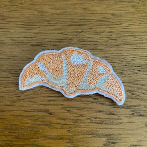 May include: Embroidered croissant-shaped patch. The patch has a light orange and white design with a white border. It is likely intended for application on clothing or accessories.
