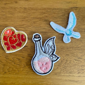May include: Three embroidered patches: a red heart with gold accents, a clear bottle with pink liquid and fairy wings, and a blue fairy with wings.