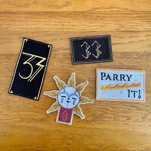 May include: A selection of embroidered patches on a wooden surface. The patches feature a black rectangle with the number "33", a rectangular patch with the text "33", a sun design, and a patch with the text "PARRY IT!".