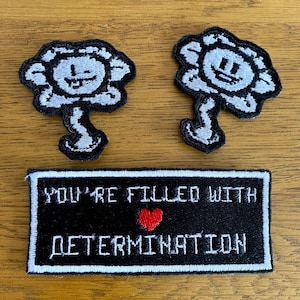 May include: Two embroidered patches with a smiling flower design, white petals and black outlines. Below, a rectangular patch with the text "YOU'RE FILLED WITH DETERMINATION" and a red heart, all in white on a black background.