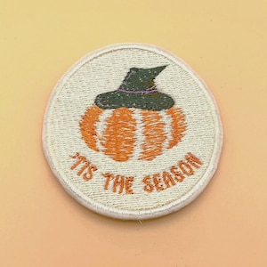 May include: A circular embroidered patch featuring an orange pumpkin wearing a dark green witch hat. The background of the patch is off-white with the text 'TIS THE SEASON' stitched in orange thread along the bottom curve of the circular design.